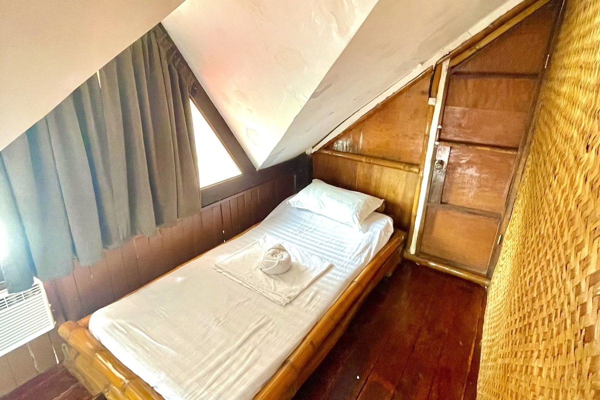 A single bed with a wooden frame and a white mattress, positioned under a slanted roof with a woven wall on one side.