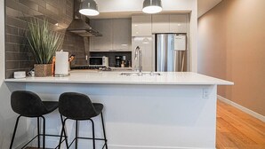 Una cocina moderna con una isla central, dos taburetes de bar, un refrigerador de acero inoxidable e iluminación colgante.