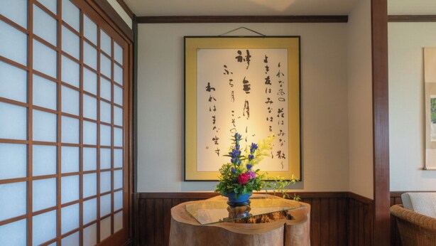 A traditional Japanese interior with tatami flooring, a wooden table with a vase of flowers, and a framed calligraphy piece on the wall. - Isoarukinoyuyado Ushiogumo (Kawazu)