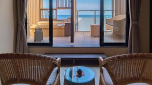 Una habitación con un ventanal grande que da a una terraza y al mar. Hay sillones de mimbre y una mesita en primer plano.