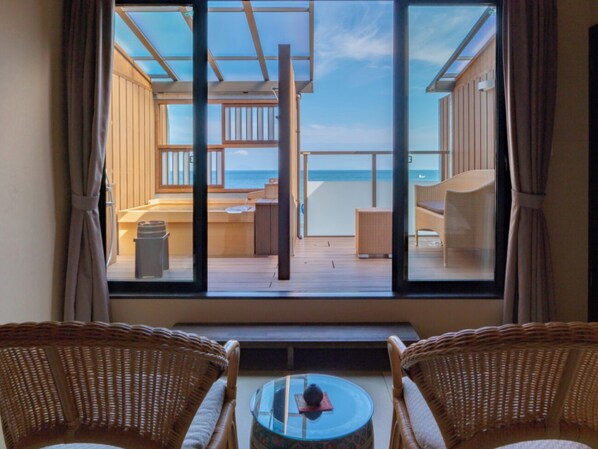 Une pièce dotée d’une grande fenêtre donnant sur une terrasse et la mer. On y trouve des fauteuils en osier et une petite table au premier plan.