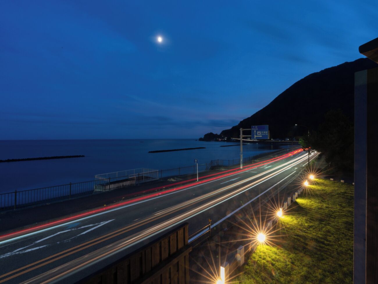Eine Küstenstraße im Dämmerlicht mit Lichtspuren von Fahrzeugen, einem klaren Himmel und einem Gebäude in der Ferne.