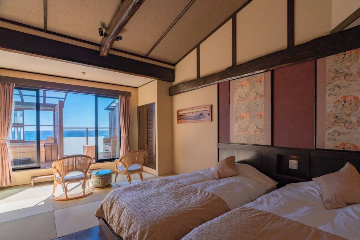 A hotel room with two beds, a balcony with wicker chairs, and a large window offering a view.
