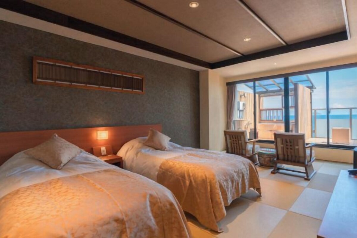 Habitación de hotel con dos camas, un ventanal con vistas, cabecero de madera y un pequeño rincón de descanso con una mesa y sillas.