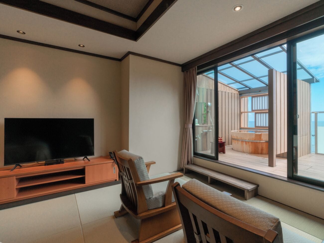 A modern living room with a flat-screen TV, wooden furniture, and a large window offering a view of an outdoor deck.