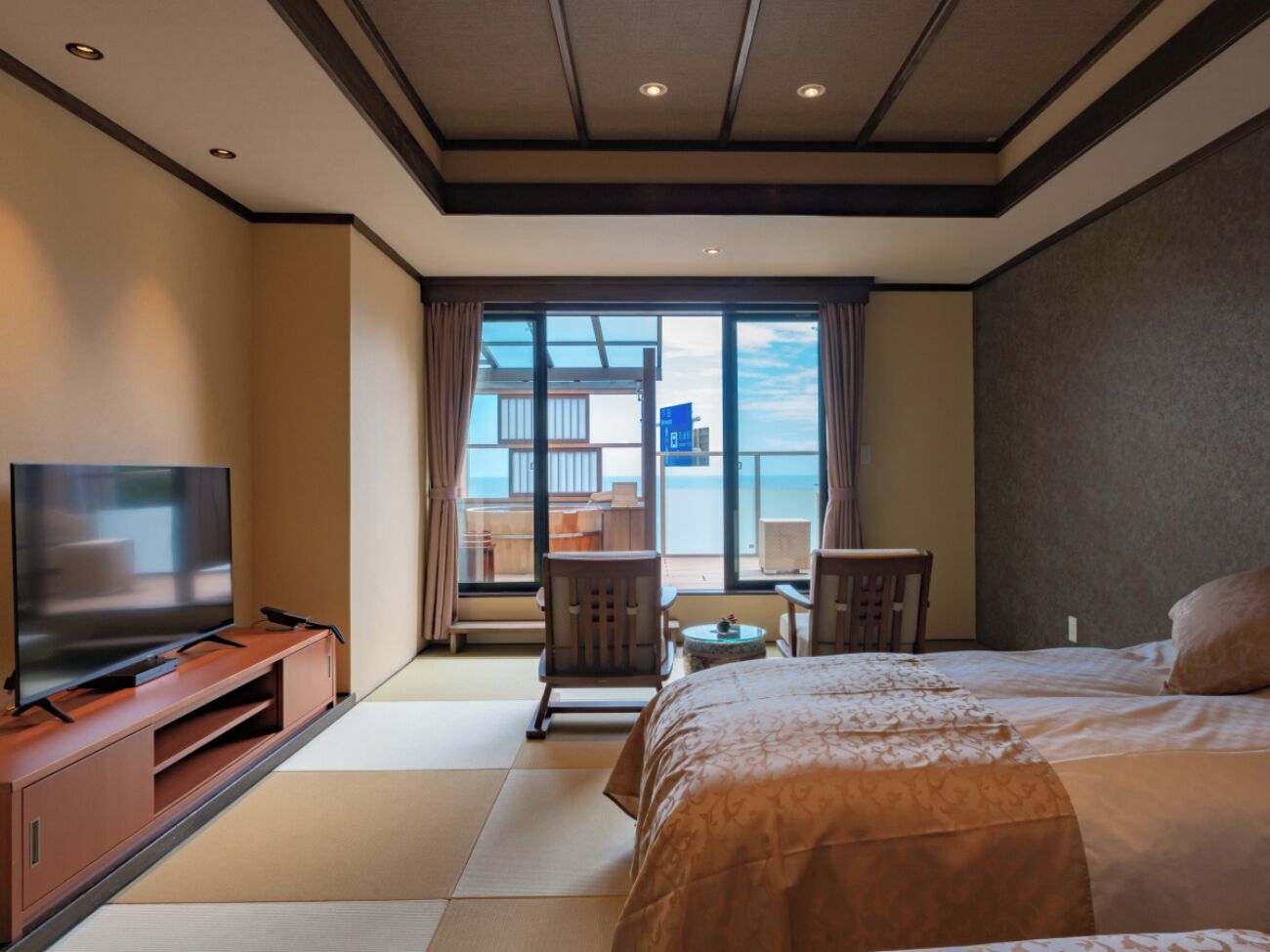 Een hotelkamer met een bed, een televisie op een houten standaard, een balkon met stoelen en uitzicht op zee.