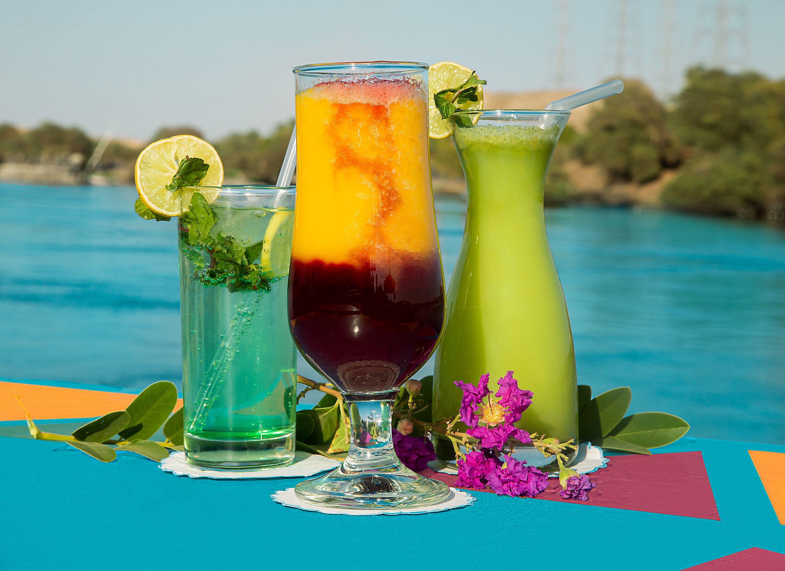 Tres bebidas coloridas con adornos, sobre una mesa cerca del agua.
