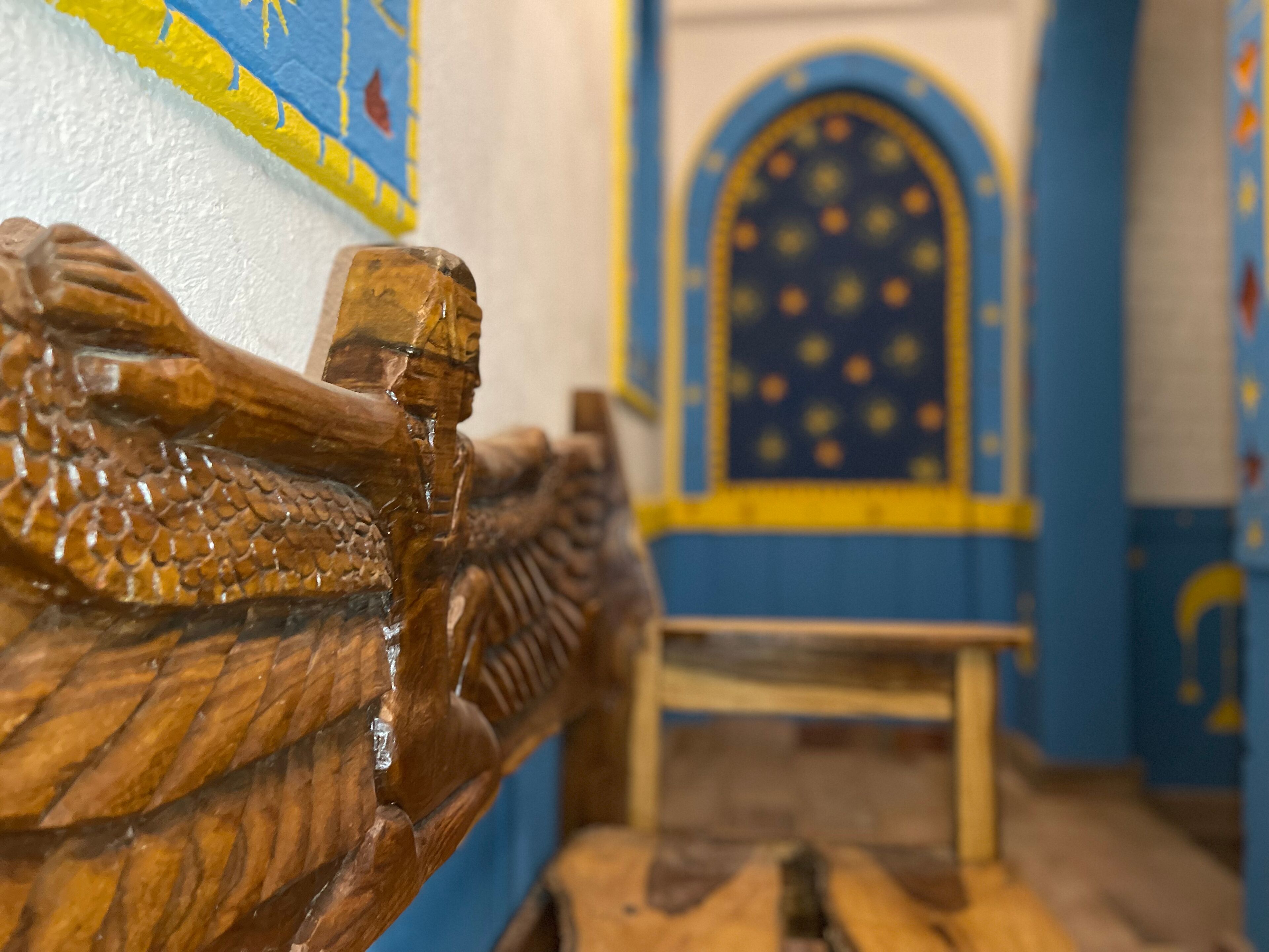 A wooden bench with a carved headrest in a room with blue and yellow patterned walls.