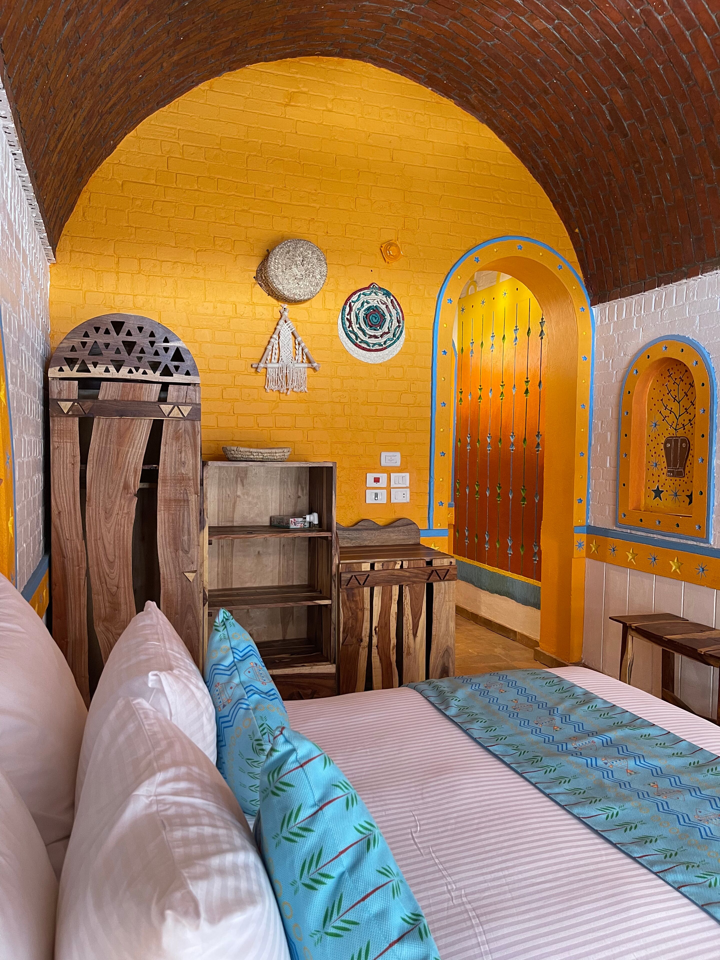 A room with a wooden door, a bed with pillows, a small wooden table, and a wall with a yellow brick pattern and decorative items.