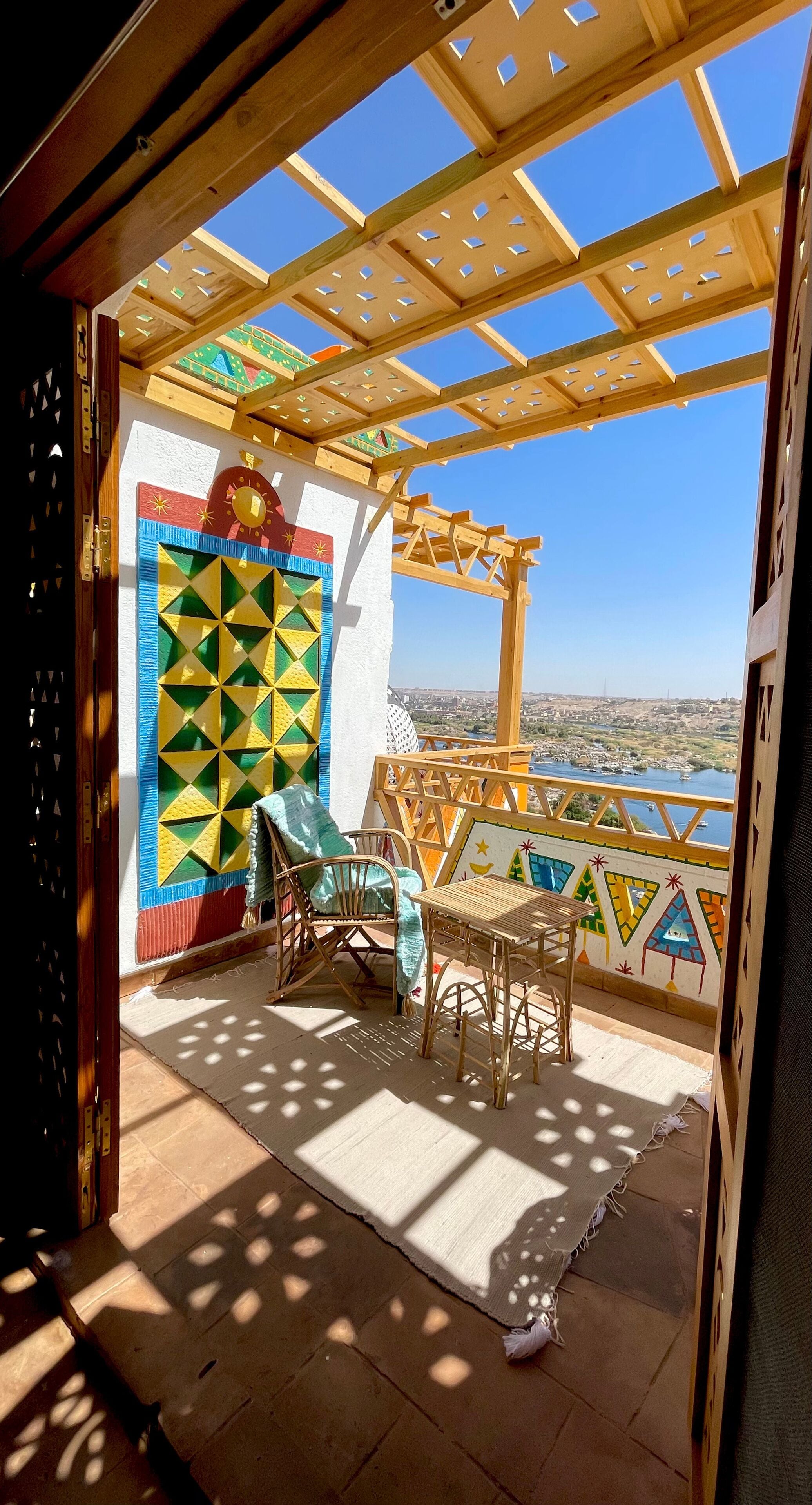 Uma varanda com pérgola de madeira, parede colorida com padrão geométrico, uma cadeira, uma mesa e vista para um rio e paisagem distante.