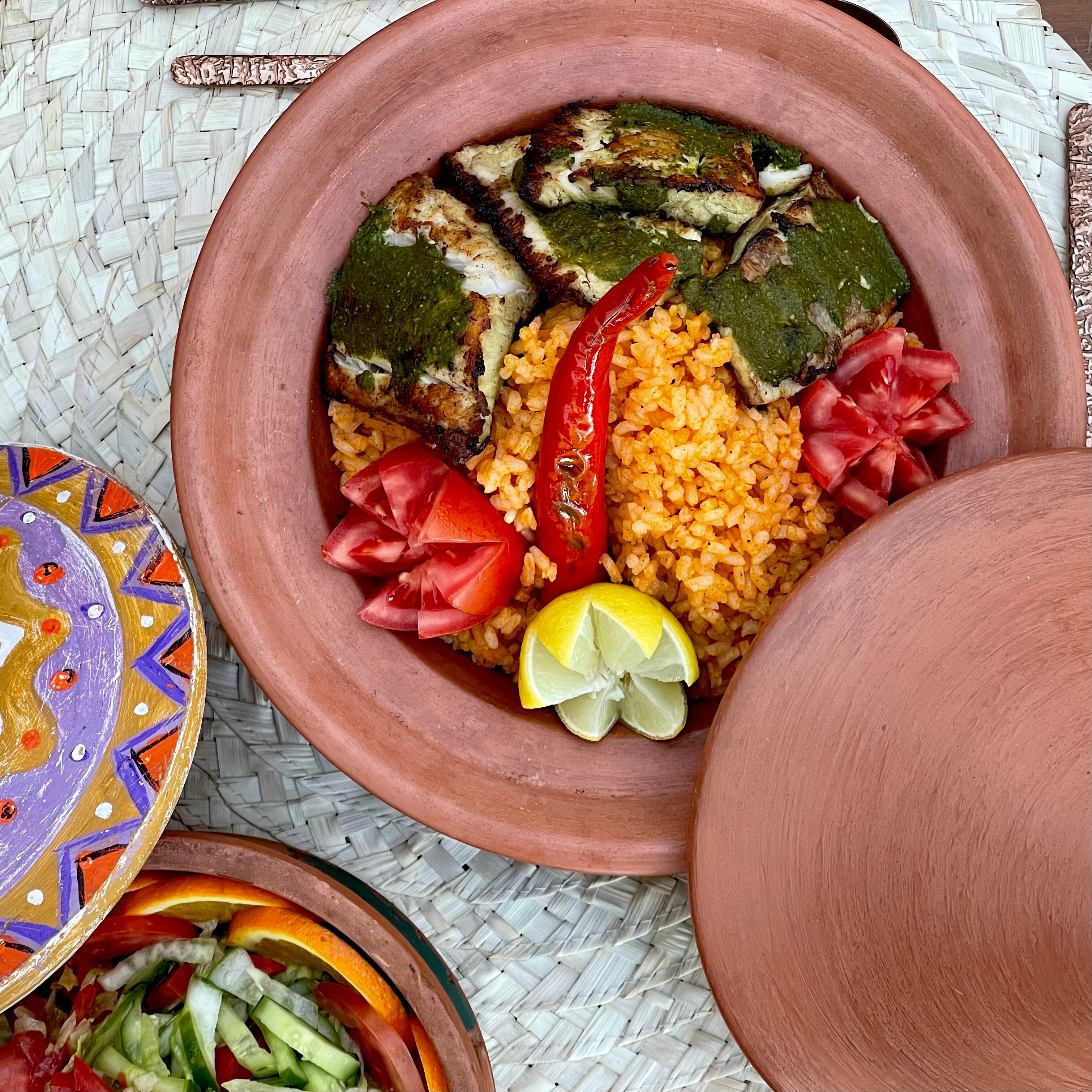 A traditional clay pot filled with rice, vegetables, and fish, accompanied by a decorated plate and a woven mat.