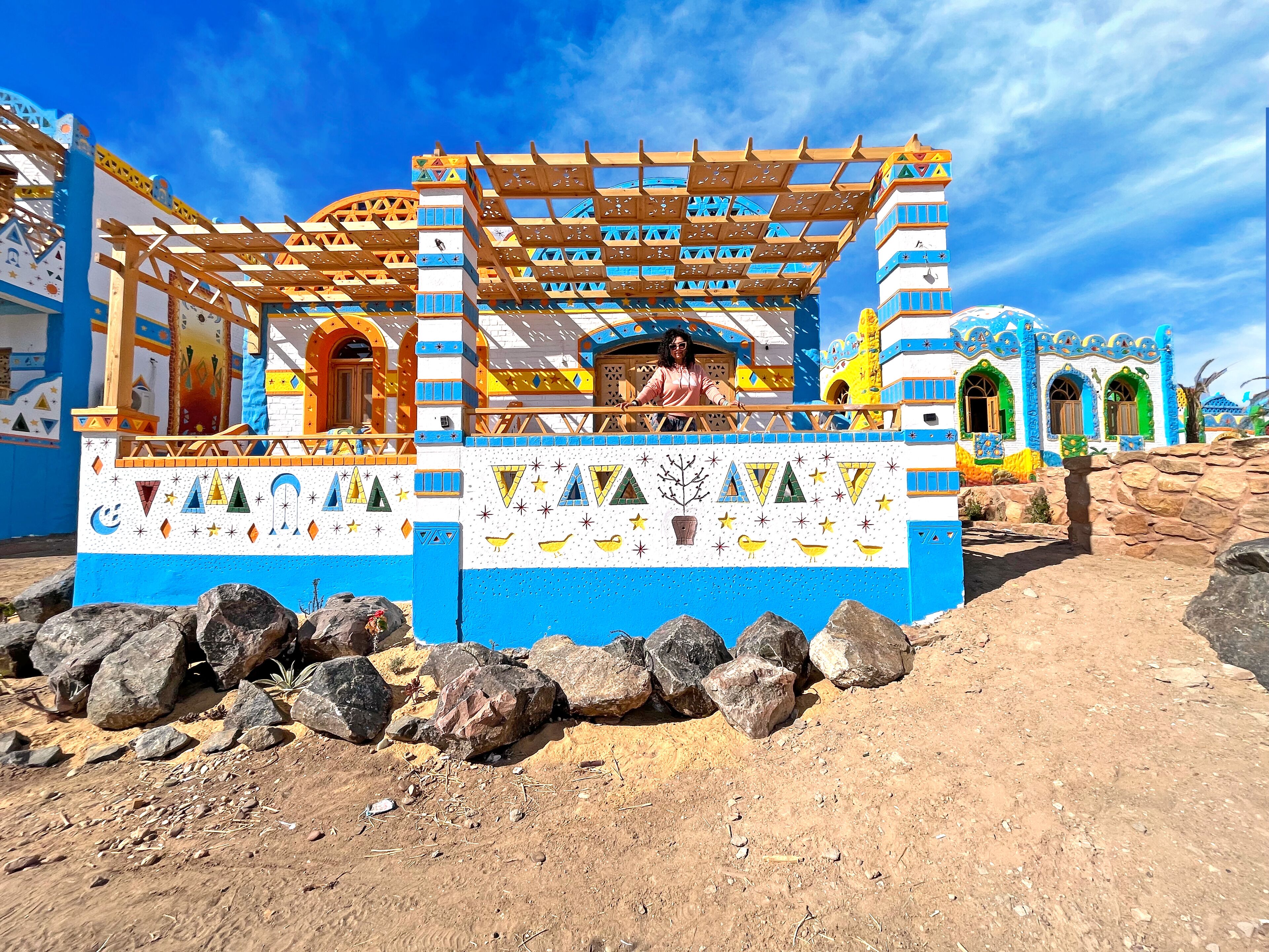 A colorful building with a blue and white facade, a wooden pergola, and a stone wall in front.