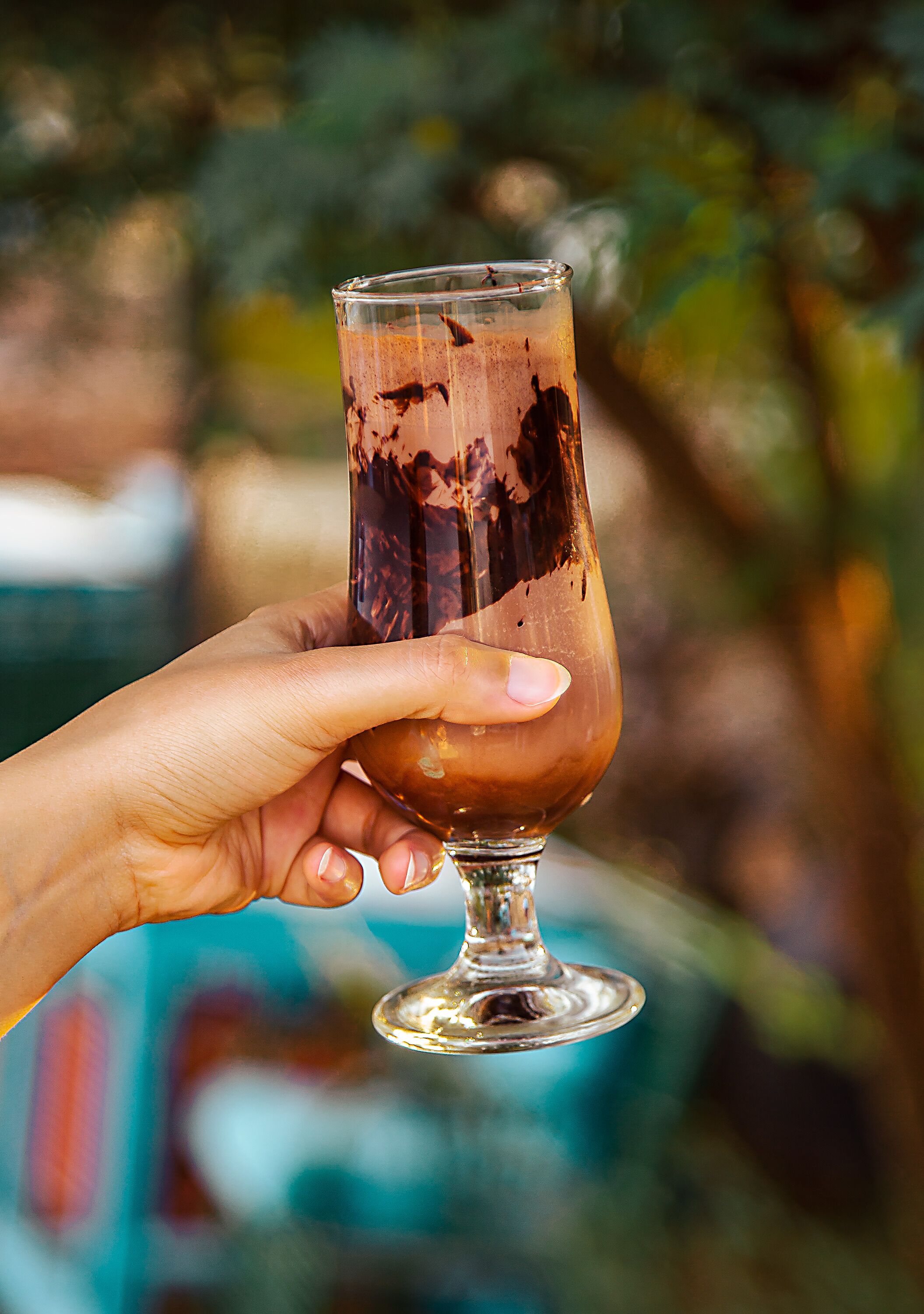 A hand holding a glass of iced coffee with a blurred background of greenery.
