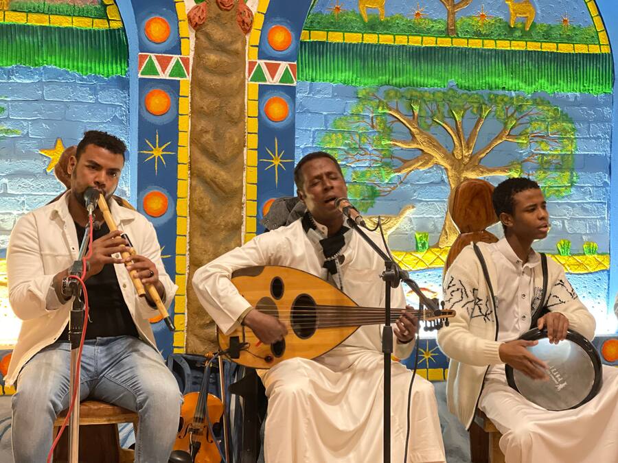 Um grupo de músicos se apresentando em um palco, com um mural colorido ao fundo.