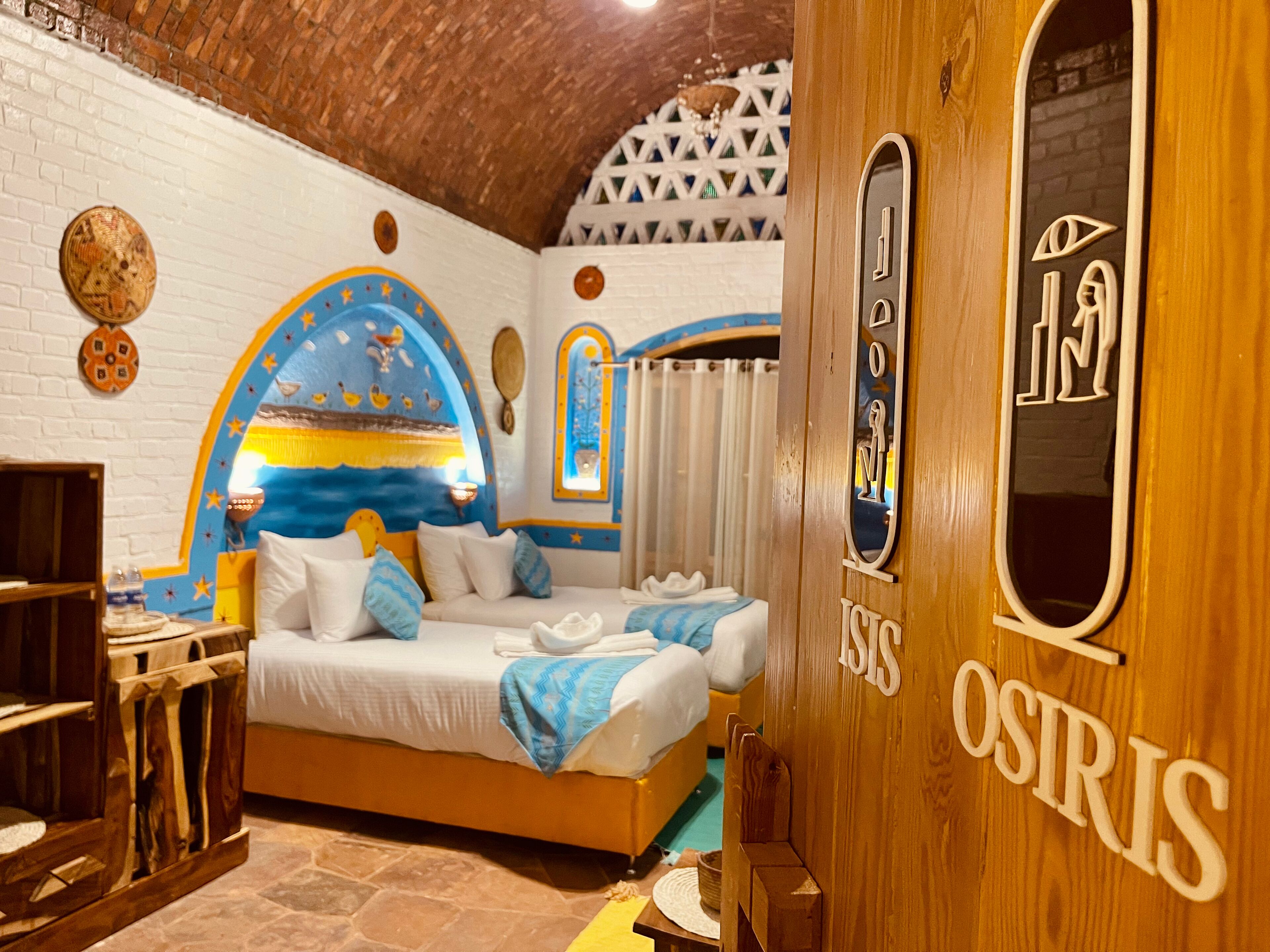 A room with a bed, a wooden door with the name 'OSIRIS', and a decorative wall with a blue and yellow mural.