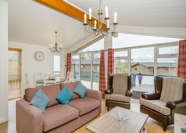 A cozy living room with a sofa, armchairs, a chandelier, a dining table, and a large window with curtains.