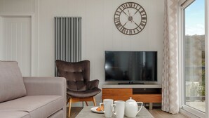A living room with a sofa, armchair, flat-screen TV, coffee table with croissants, and a wall clock. - 3 bedroom accommodation in Cheddar (Cheddar)