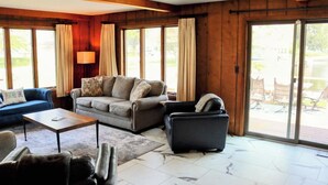 A spacious living room with a sofa, armchairs, and a coffee table. Large windows provide natural light and a view of the outdoors. - 4B Lakefront home off M22 with access to Lake Michigan! (Onekama)