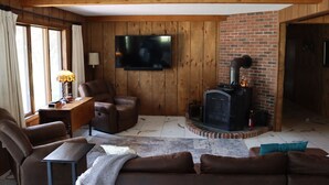 A cozy living room with a fireplace, leather sofas, a television, and wooden paneling. - 4B Lakefront home off M22 with access to Lake Michigan! (Onekama)
