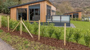 A modern wooden cabin with large windows, a deck, and a view of a hilly landscape. - 4 bedroom accommodation in Cheddar (Cheddar)