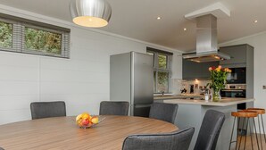 A modern kitchen with a central island, a dining area with a round table, and grey upholstered chairs. - 4 bedroom accommodation in Cheddar (Cheddar)