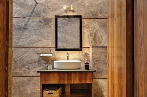 Un baño con pared de mármol, tocador de madera, lavamanos redondo, espejo e iluminación.