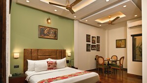 A hotel room with a large bed, a dining table with chairs, a ceiling fan, and wall-mounted artwork. - Hotel Sethi Legacy (Haridwar)