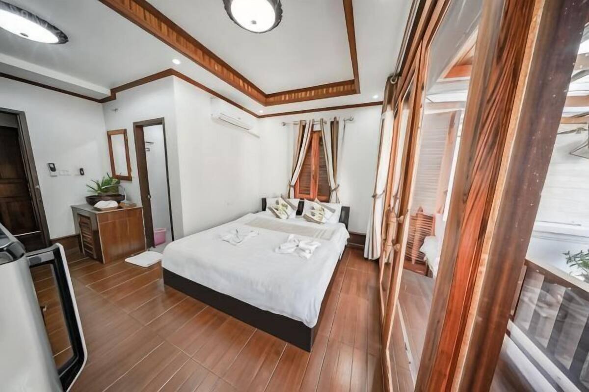 A bedroom with a large bed, wooden flooring, a ceiling light, a bedside table, and a window with curtains.