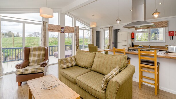 A spacious living room with a sofa, a chair, a wooden table, and a kitchen area with a stove and refrigerator. - Wordsworth Premier Select (Bewaldeth, Keswick)