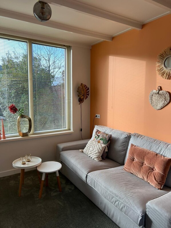 Uma sala de estar com um sofá cinzento, duas mesinhas redondas, um painel decorativo em forma de folha e uma janela com persianas.