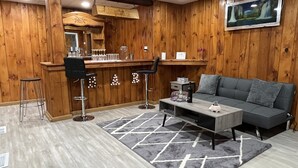 A room with wooden walls, a bar area with stools, a grey sofa, and a patterned rug.