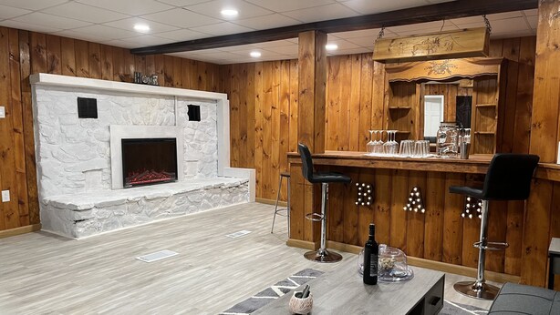 A modern living room with a stone fireplace, wooden panel walls, a bar area with stools, and a patterned rug.