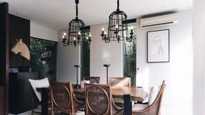 A dining area with a wooden table and wicker chairs, black pendant lights, and a framed picture on the wall. - Villa Ambergris by Nakula (Nusa Dua)