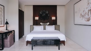 A hotel room with a large bed, two bedside lamps, a headboard with a decorative panel, a small table with a lamp, and a bench. - Villa Ambergris by Nakula (Nusa Dua)