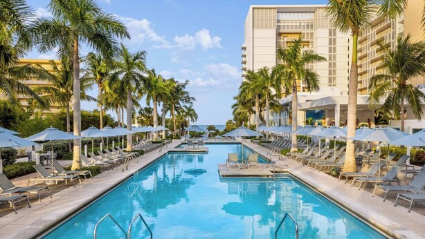 A swimming pool with lounge chairs and umbrellas, surrounded by palm trees and a hotel building.