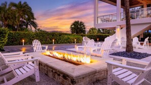 A fire feature surrounded by white Adirondack chairs, a pool, and a modern building with large windows. - 3BR Poolside! Marriott Crystal Shores Resort - Luxurious Condo - Beachfront Resort (Marco Island)