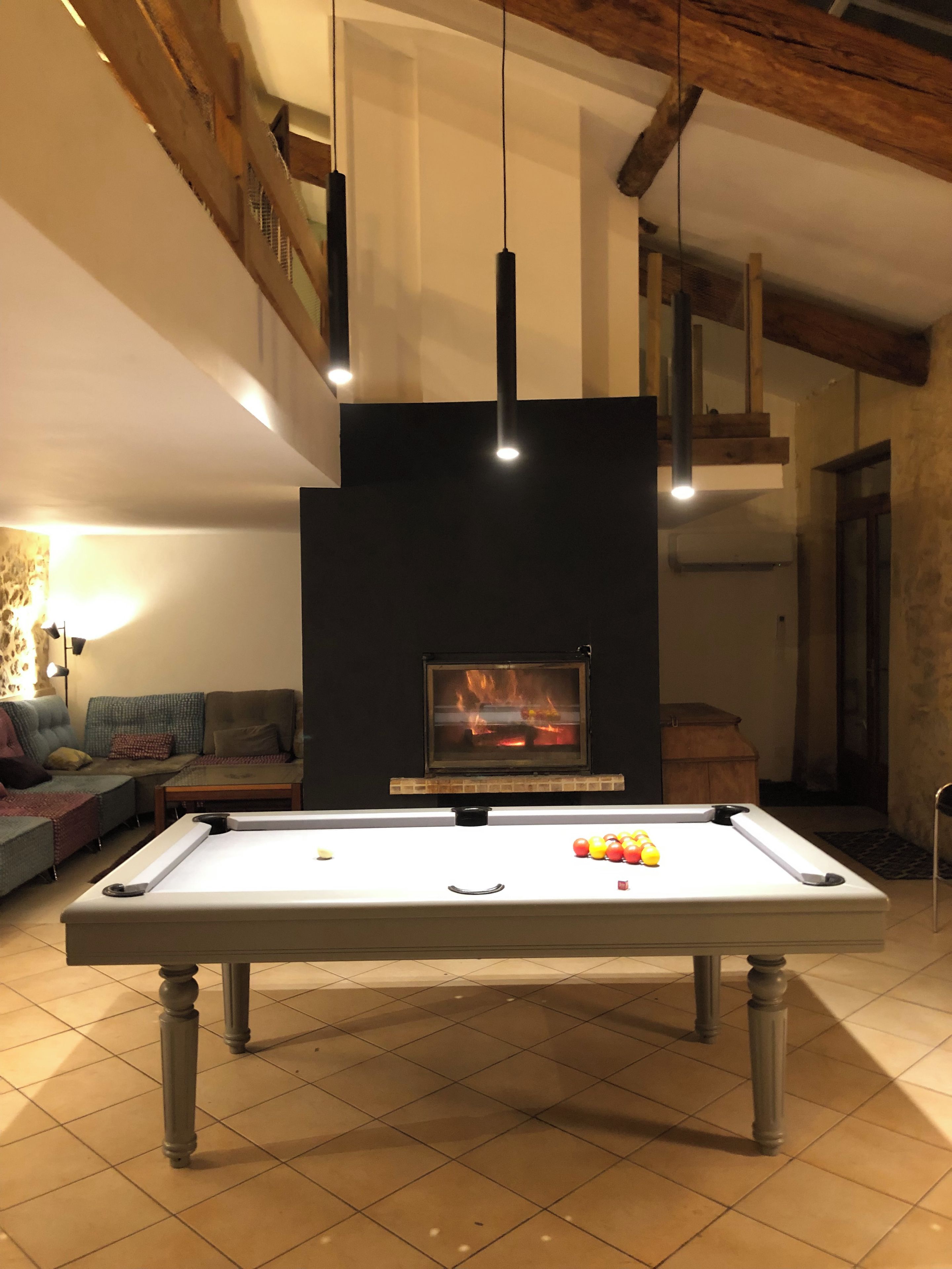 A pool table with balls, a fireplace, and a sofa in a room with wooden beams and hanging lights.