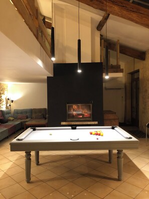 Une table de billard avec des billes, une cheminée et un canapé dans une pièce aux poutres apparentes et aux luminaires suspendus.