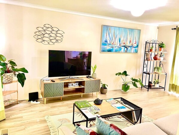 A living room with a TV, a painting, a plant shelf, and a coffee table. - Corner Cottage (Dandenong North)