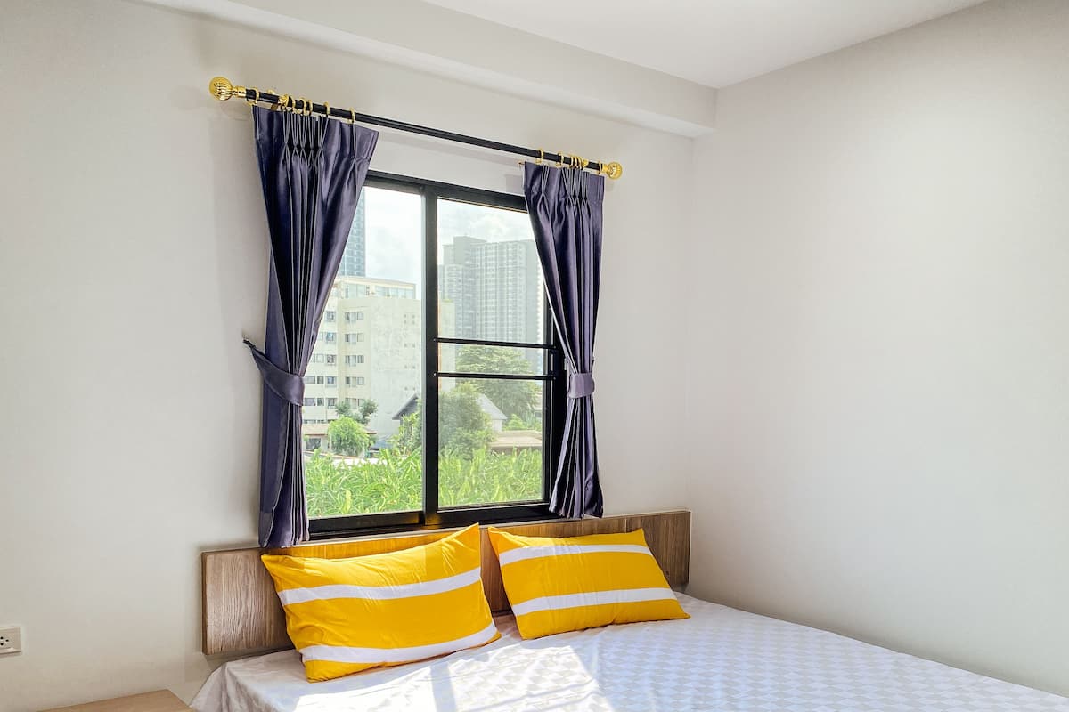 A bedroom with a bed, two pillows, a nightstand, a window with curtains, and a view of buildings and greenery outside.