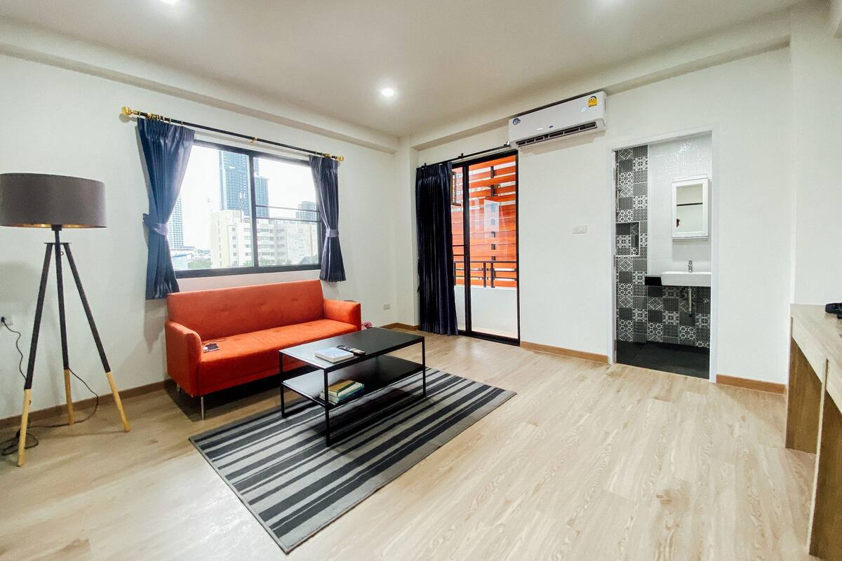A modern living room with a red sofa, a black coffee table, and a floor lamp. There is a large window with curtains, an air conditioning unit, and a view of buildings outside.