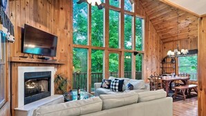 A cozy cabin interior with a fireplace, a television, a sofa, and a dining area. - 🛏️ Knotty Nice | Sleeps 11 in 2BR + Game Room + Hot Tub 🏔️ (Sevierville)