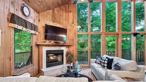 A cozy cabin interior with a fireplace, a television, and a large window overlooking greenery. - 🛏️ Knotty Nice | Sleeps 11 in 2BR + Game Room + Hot Tub 🏔️ (Sevierville)