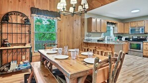 A wooden dining area with a table set for four, a chandelier, and a view of the outdoors through a window. - 🛏️ Knotty Nice | Sleeps 11 in 2BR + Game Room + Hot Tub 🏔️ (Sevierville)