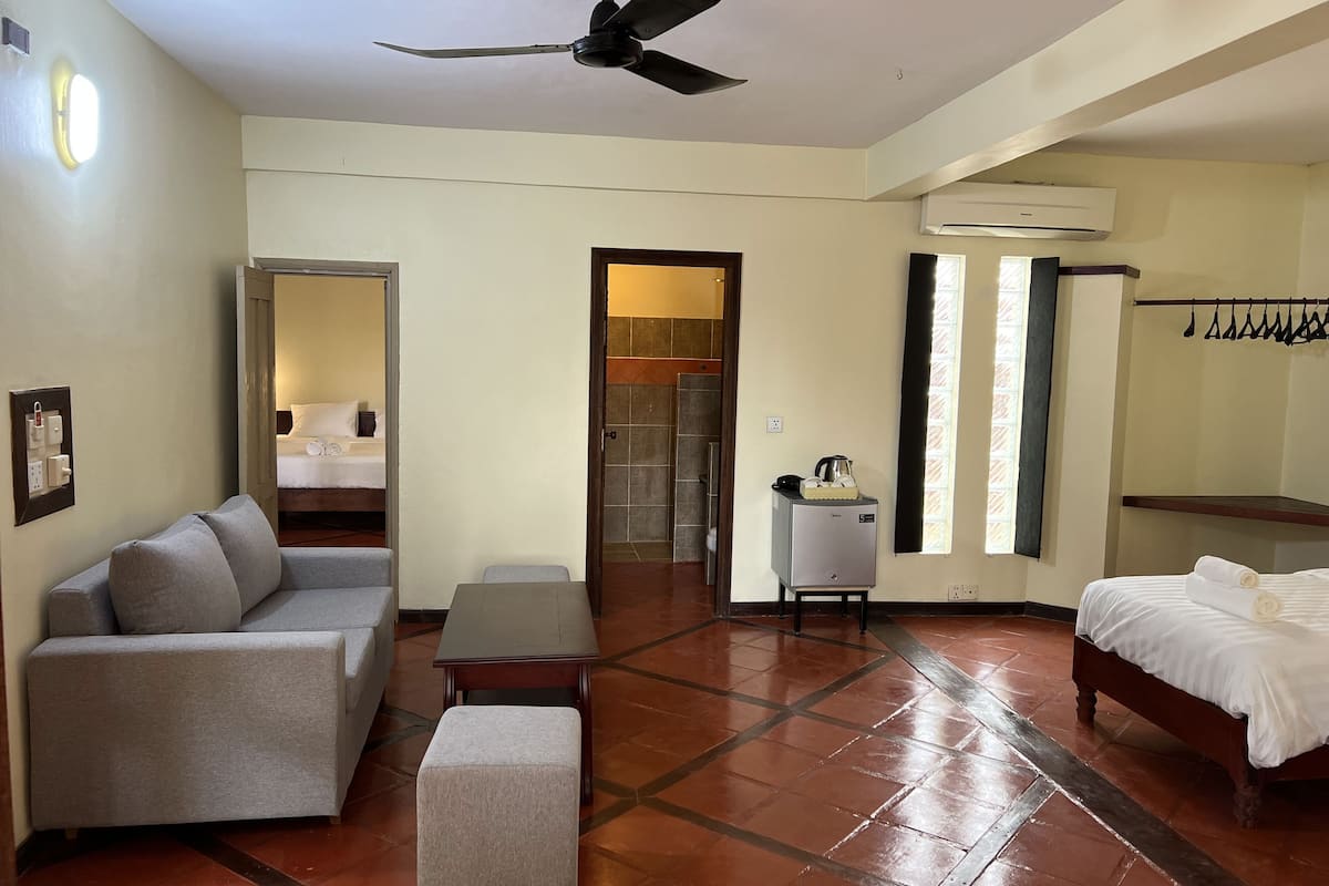 A hotel room with a sofa, a small table, a bed, a ceiling fan, an air conditioning unit, and a window with curtains.