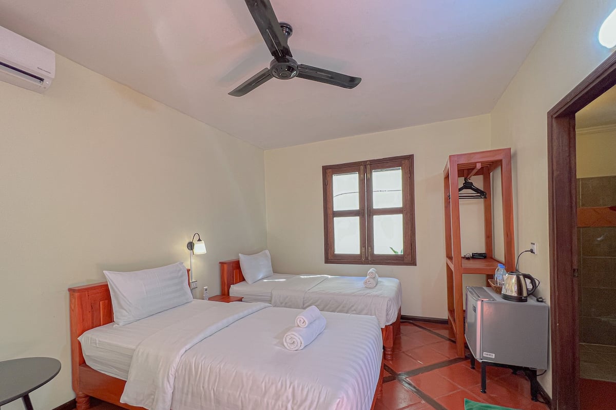 A hotel room with two beds, a ceiling fan, a window, a mirror, a small table, and a kettle.