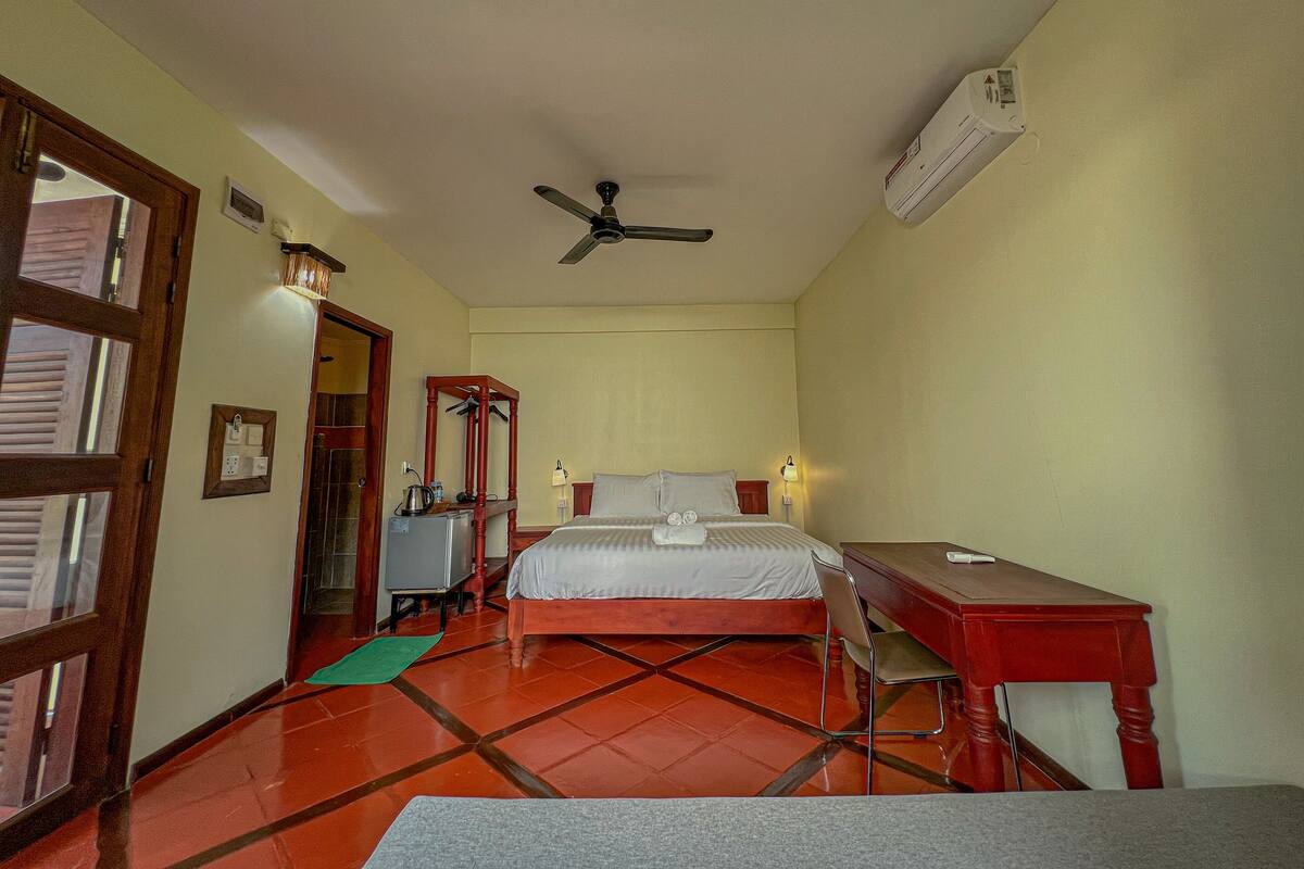 A hotel room with a bed, a desk, a chair, a ceiling fan, and an air conditioning unit.