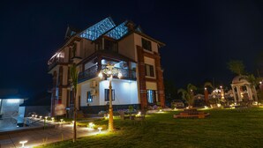 A multi-story hotel with a well-lit exterior, a swimming pool, and a garden area with outdoor seating. - Matoshree Club and Resort (Palampur)