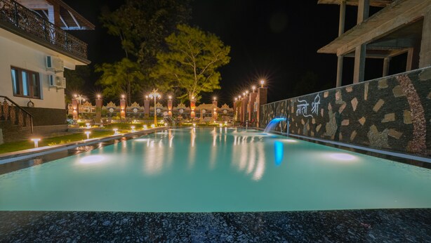 A swimming pool with a waterfall feature, surrounded by a stone wall and illuminated by lights. - Matoshree Club and Resort (Palampur)