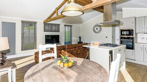 Een moderne keuken met een centraal eiland, houten balken in het plafond, een ronde eettafel met fruit en een televisie in de woonkamer.